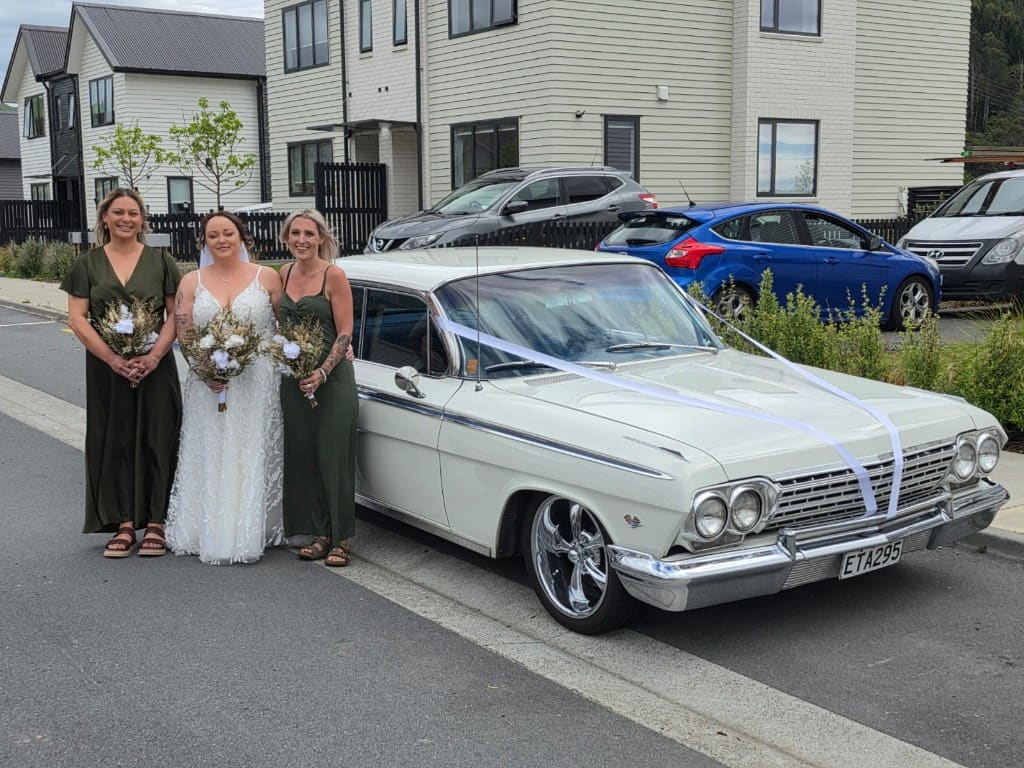 Bride and two bridesmaids with dried-flower bouquets standing beside the white Impala, wedding ribbon on the bonnet.